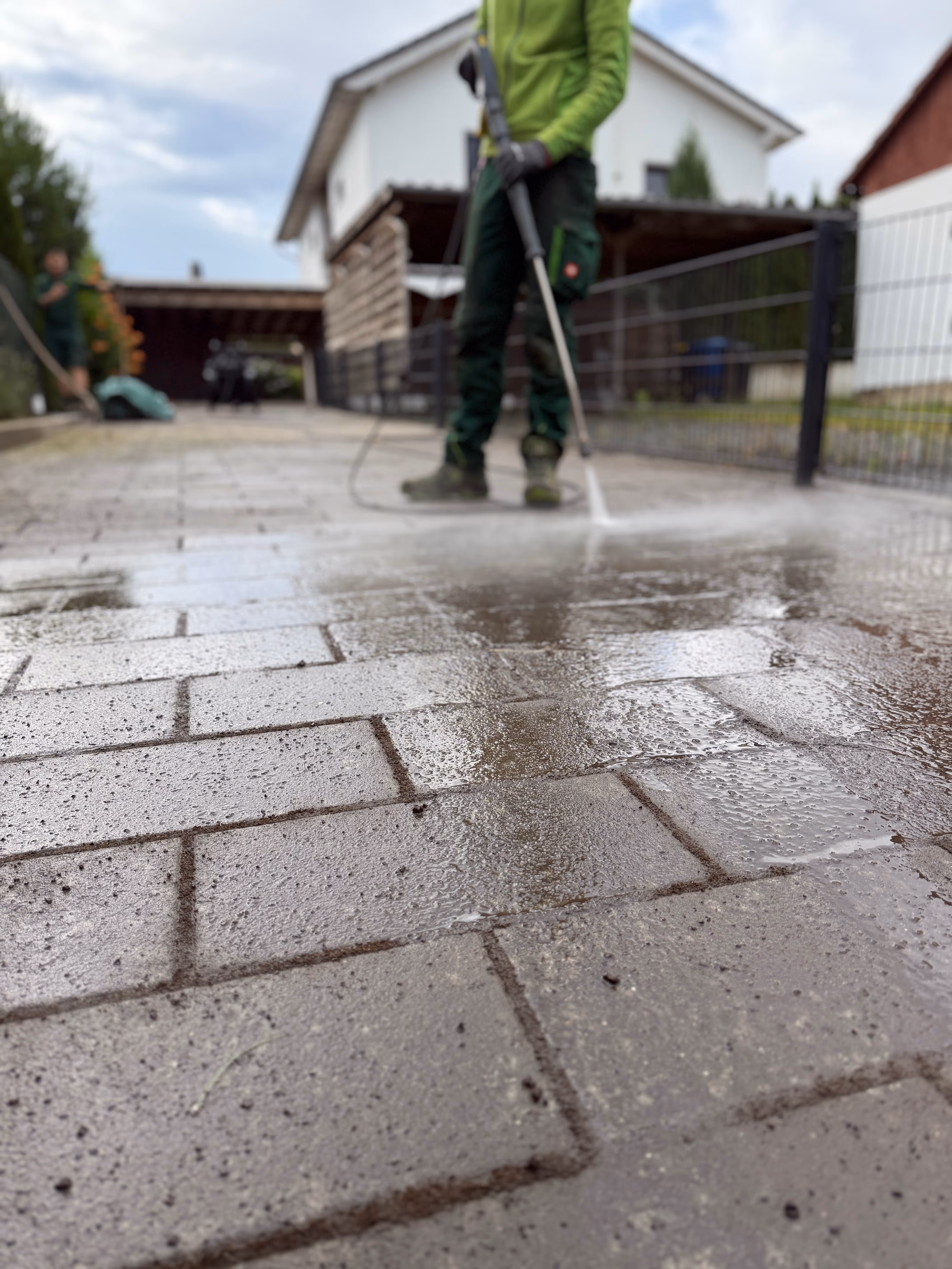 Mitarbeiter reinigt eine gepflasterte Einfahrt vor einem Wohnhaus mit dem Hochdruckreiniger
