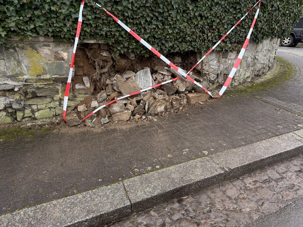 Vorher: herausgebrochene Natursteinmauer unter einer Efeuhecke, mit Flatterband abgesperrt