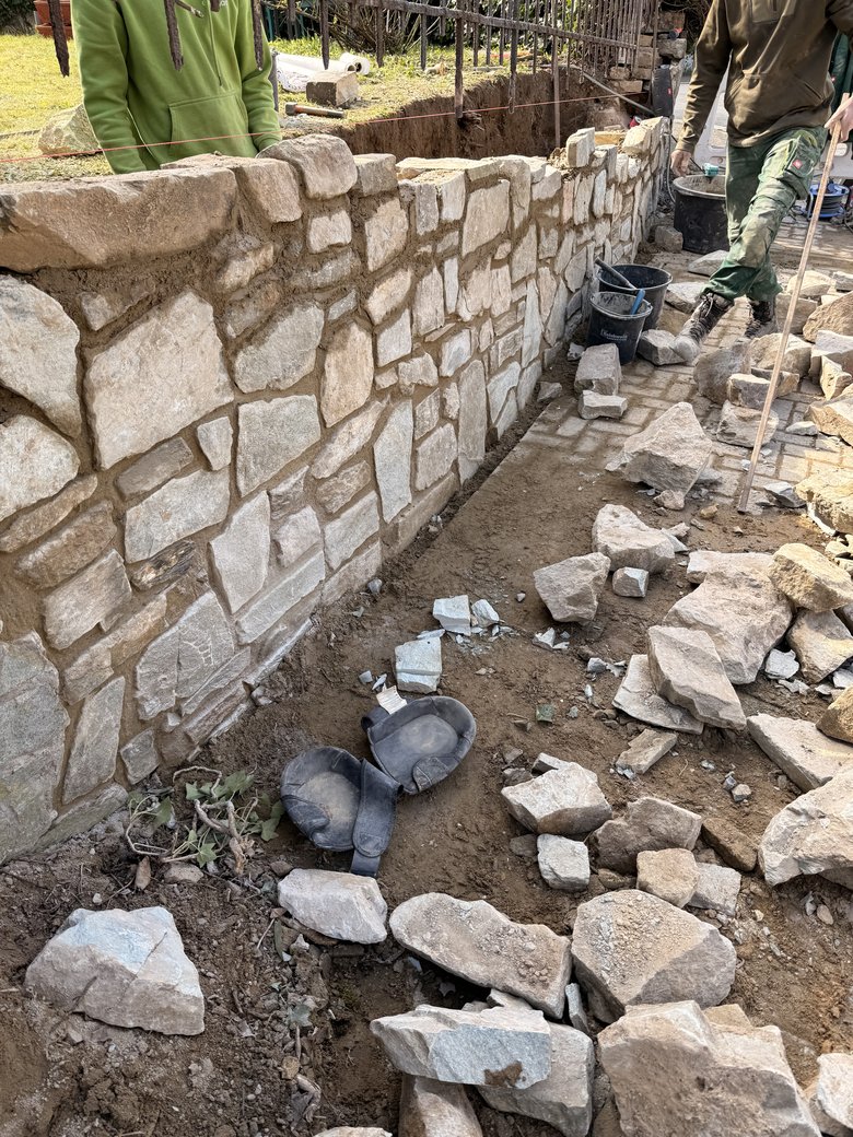 Zwei Mitarbeiter beim Wiederaufbau einer Natursteinmauer, davor sortierte Steine und Knieschoner
