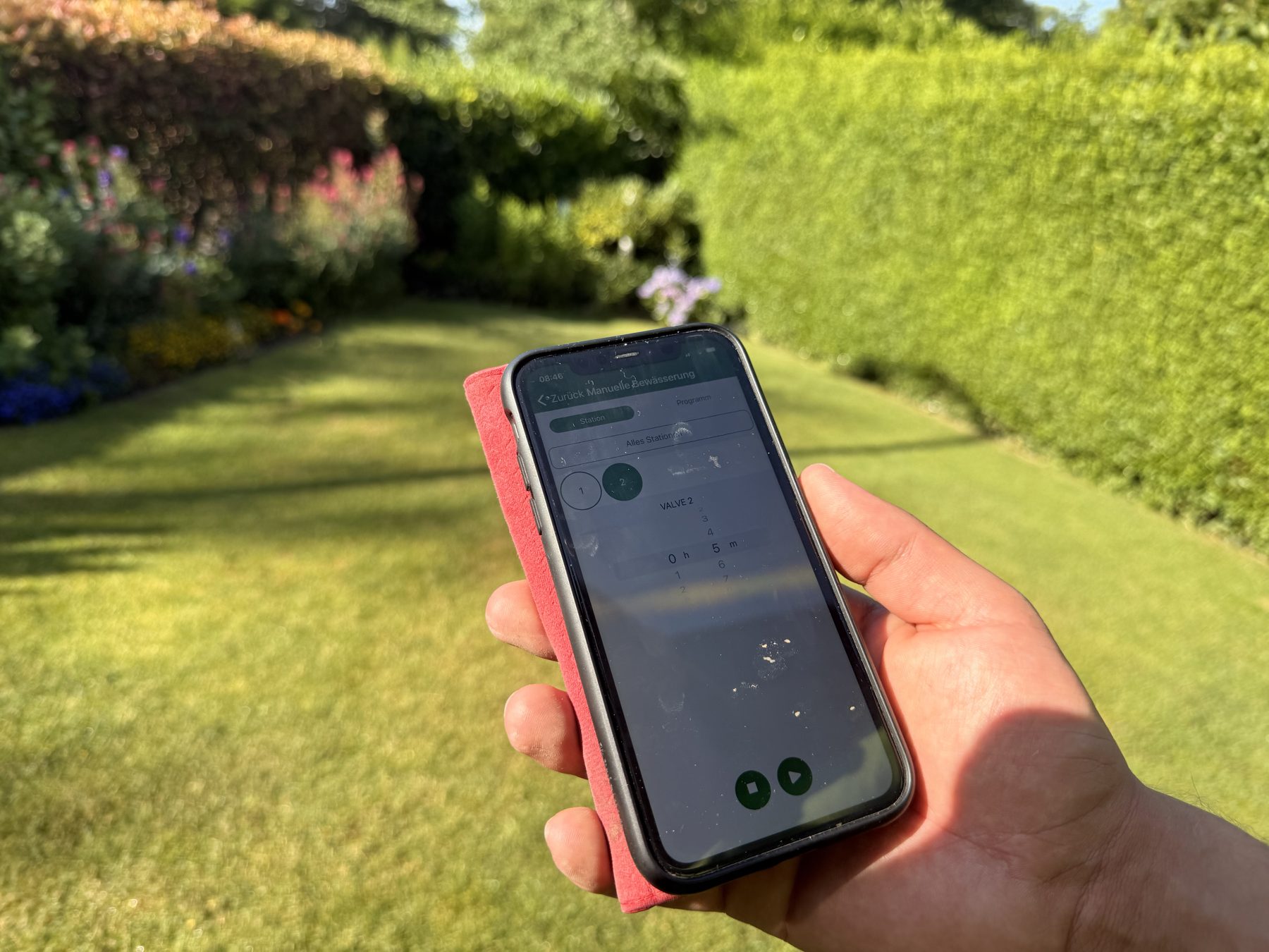 Smartphone mit Bewässerungs-App zur Steuerung einzelner Zonen, im Hintergrund Rasen und Hecke