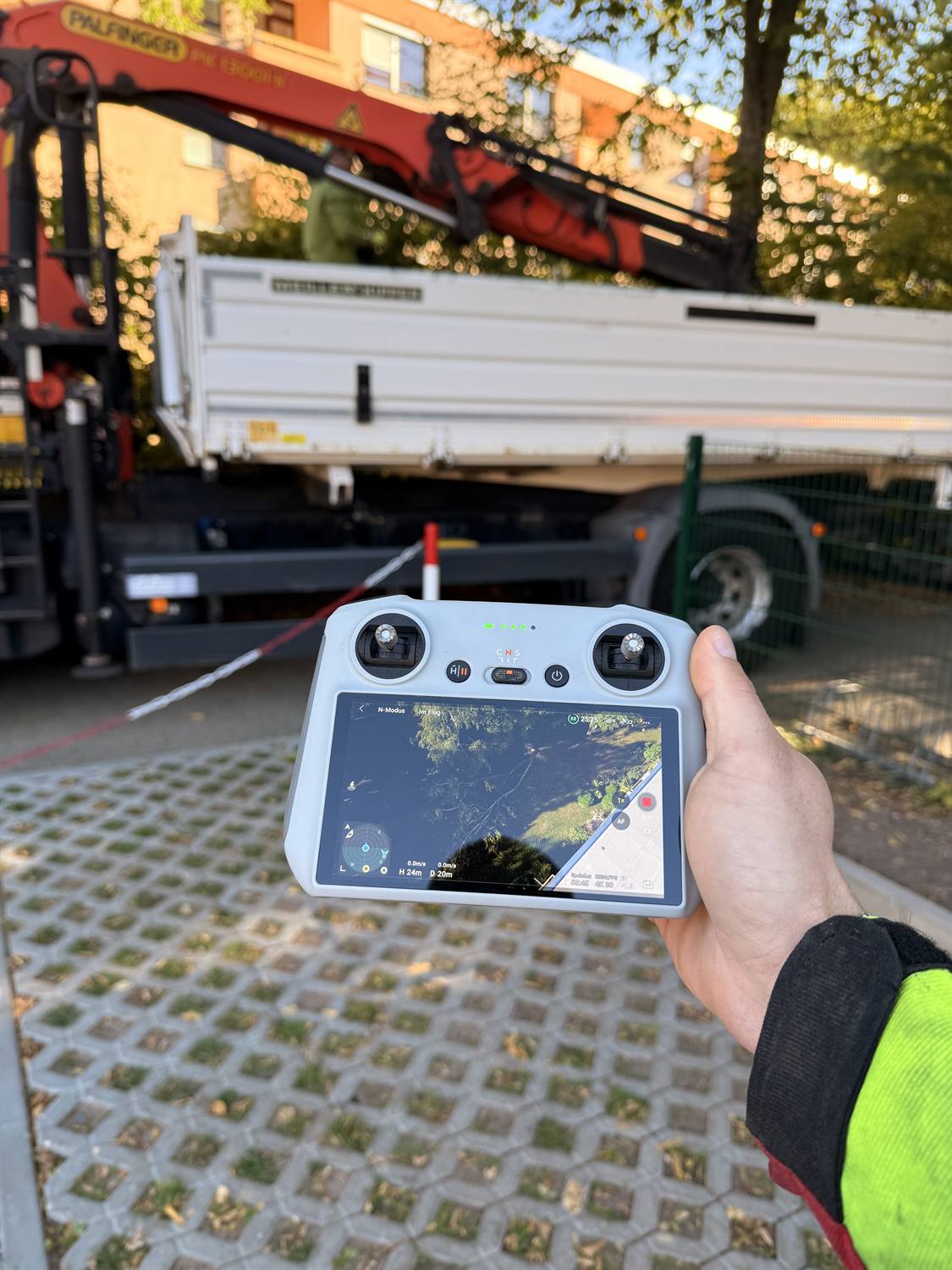 Hand hält die Fernsteuerung einer Drohne, auf dem Bildschirm die Baustelle von oben, dahinter der LKW mit Ladekran