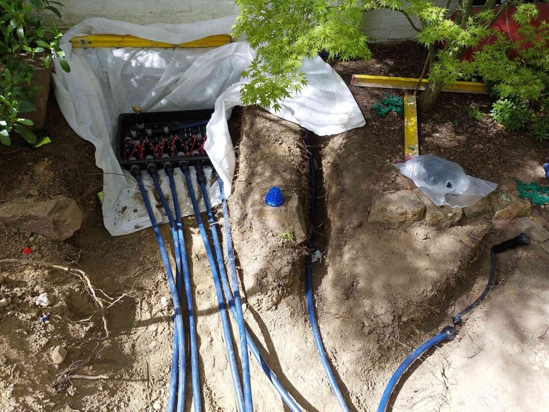 Offene Ventilbox im Beet mit sechs abgehenden blauen Leitungen, darunter weißes Vlies