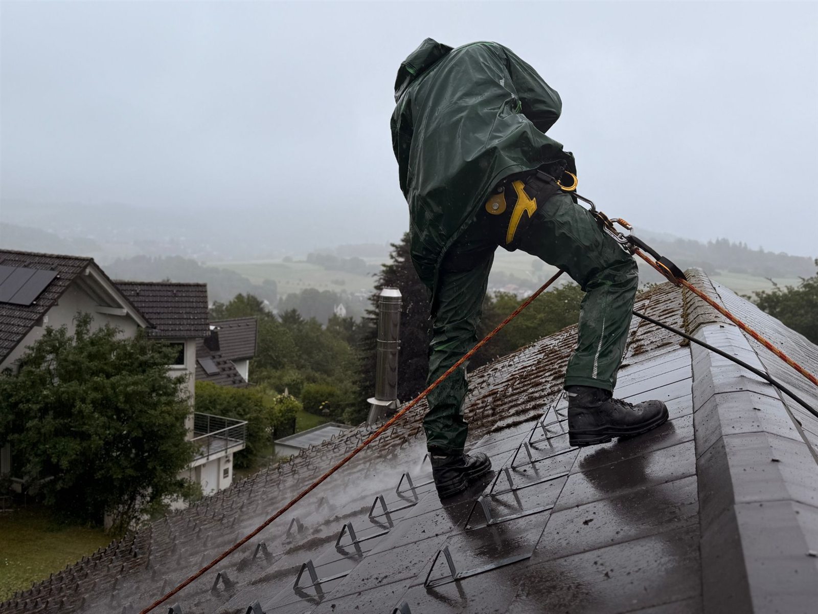 Mitarbeiter mit Auffanggurt und Seilsicherung reinigt das Dach mit dem Hochdruckreiniger, im Hintergrund das Tal im Nebel