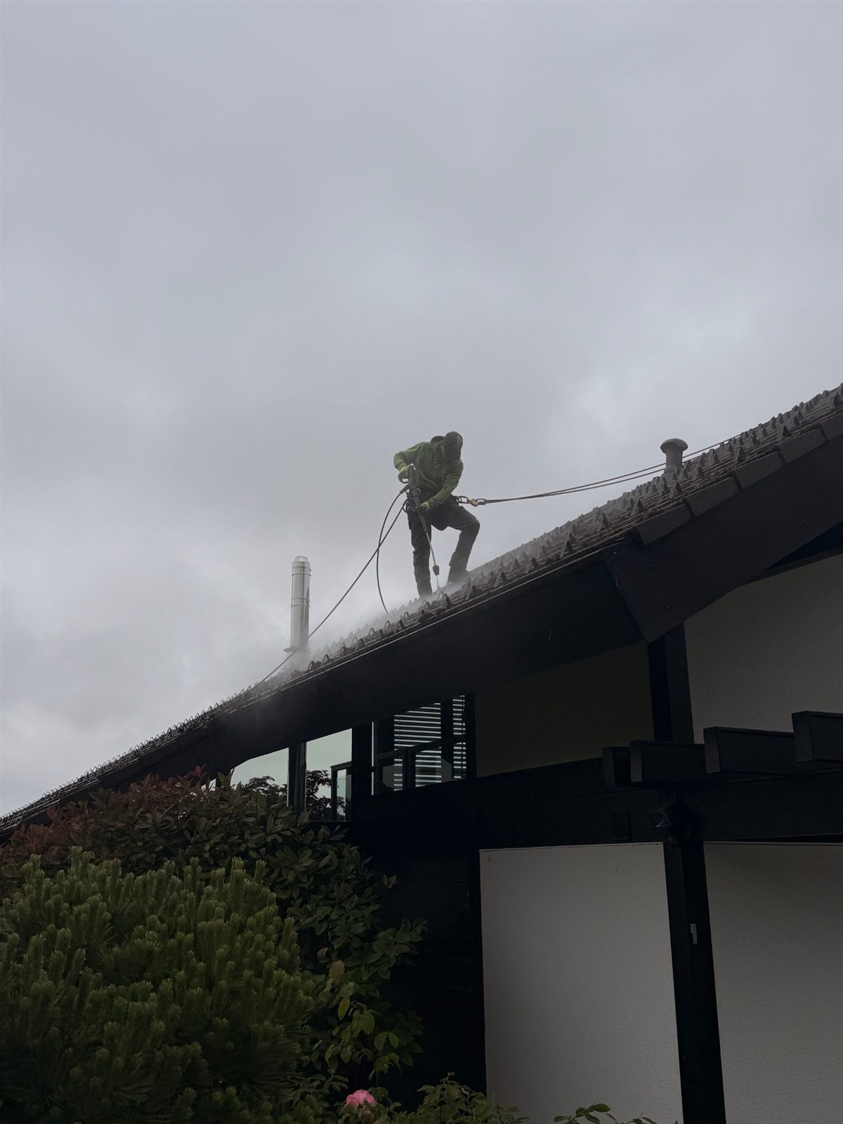 Mitarbeiter auf dem First im Sprühnebel des Hochdruckreinigers, gesichert am Seil