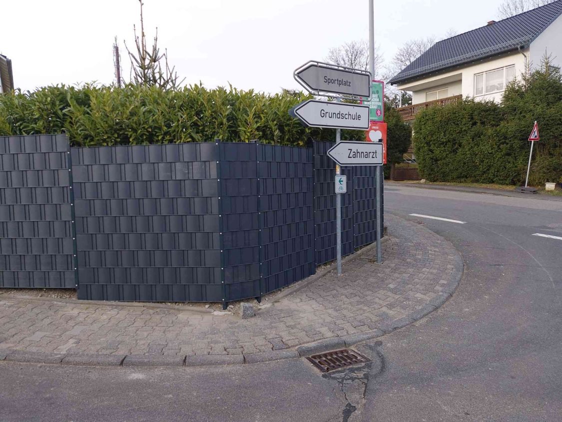 Zaun mit Sichtschutz an der Straßenecke, davor Wegweiser zu Sportplatz und Grundschule