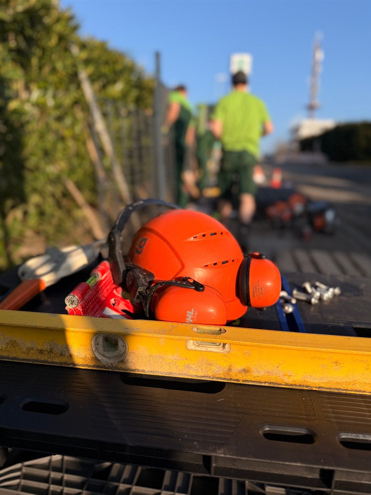 Schutzhelm mit Gehörschutz und Wasserwaage auf der Werkbank, im Hintergrund das Team am Zaun