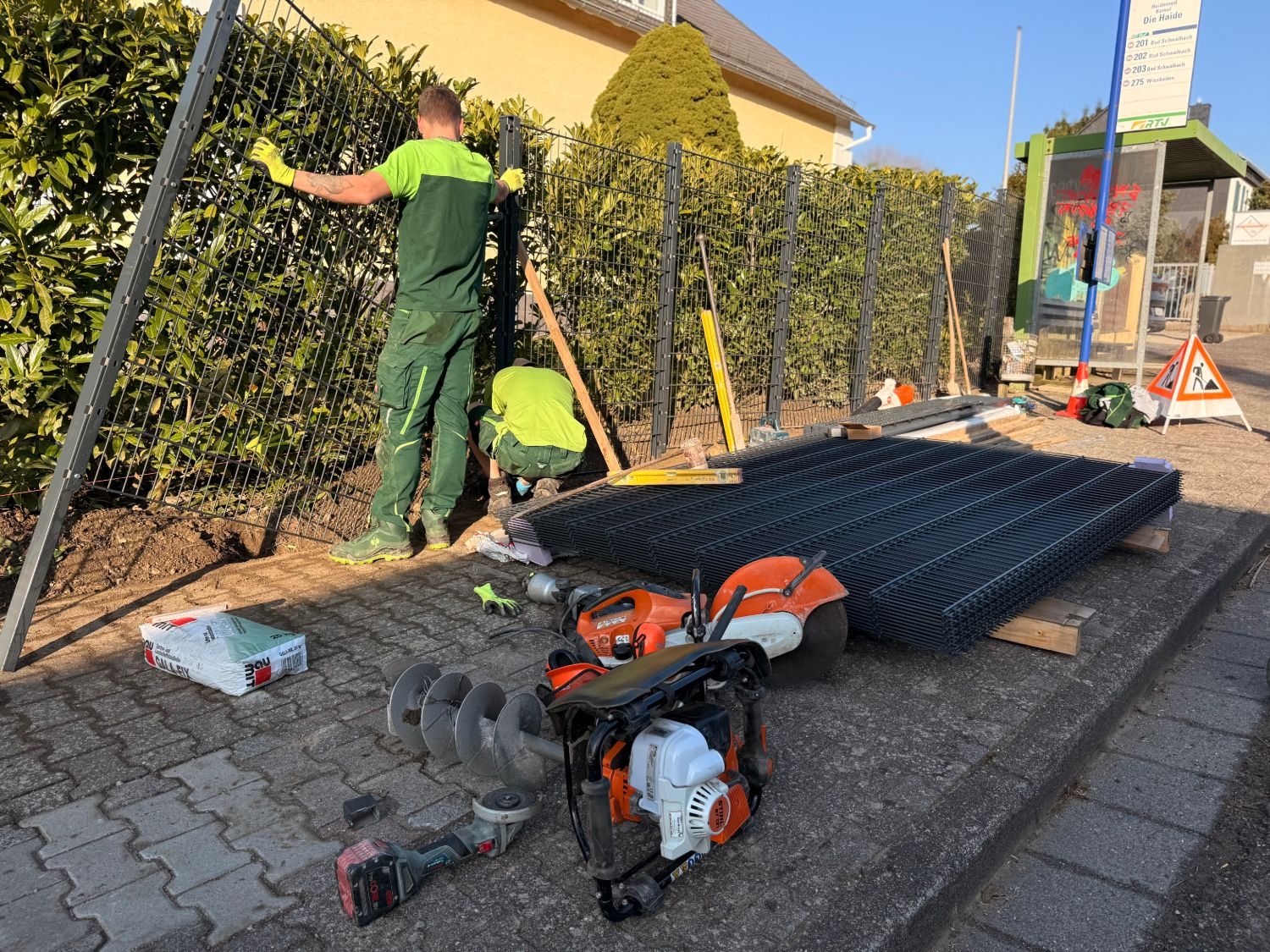 Zwei Mitarbeiter montieren die ersten Zaunfelder vor der Hecke, daneben Werkzeug und ein Stapel Matten