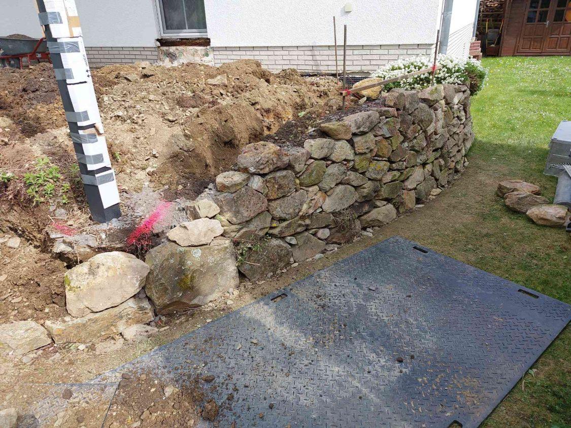Alte Trockenmauer am Rand der Baustelle, dahinter der Aushub, die Stütze der Überdachung ist mit Pappe umwickelt
