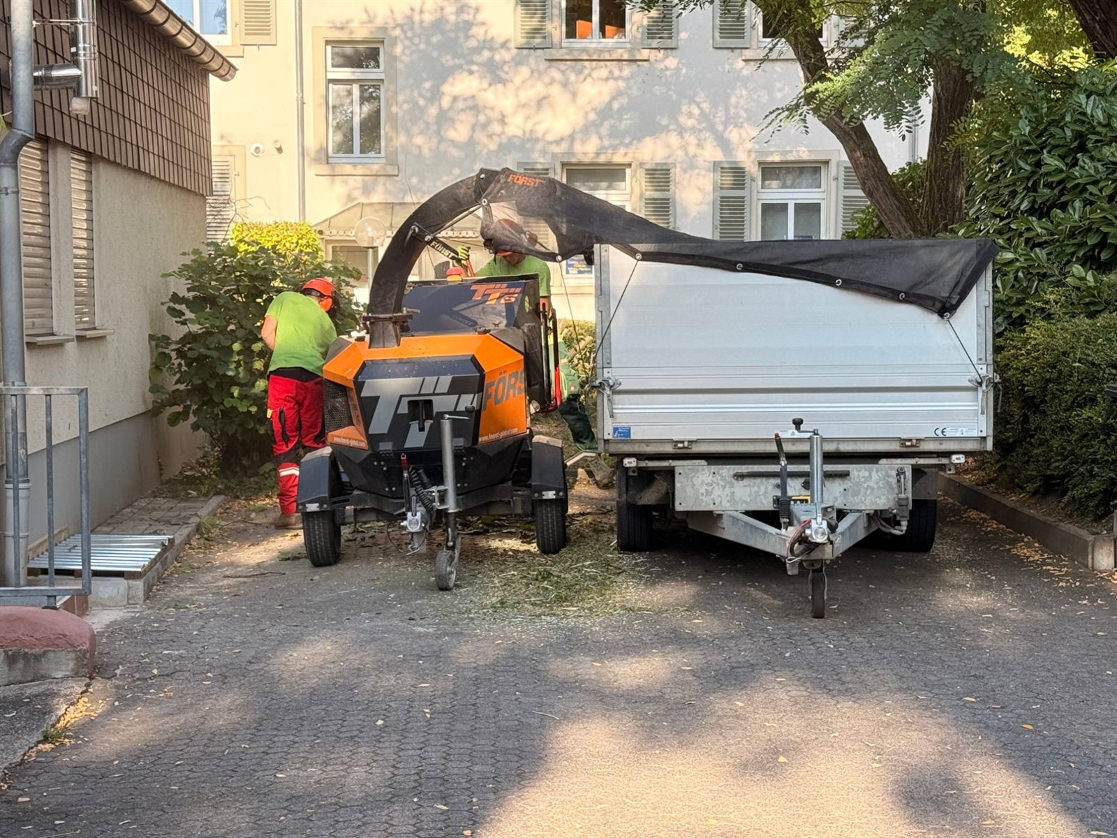 Häcksler und Anhänger in der Hofeinfahrt, ein Mitarbeiter führt Schnittgut zu