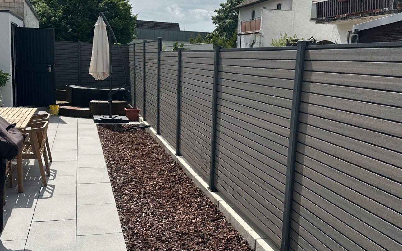 Neuer Sichtschutzzaun mit grauen Dielen und anthrazitfarbenen Pfosten entlang einer Terrasse mit Rindenmulchbeet